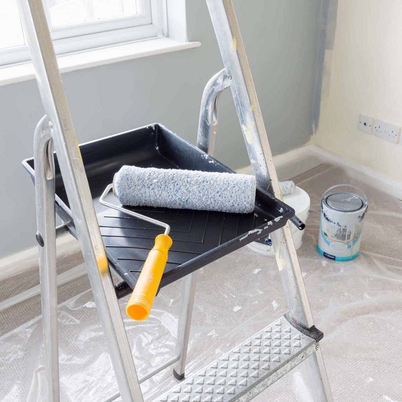 Painting and decorating with a roller and tray on a set of metal step ladders with cans of paint and a dust sheet in the background