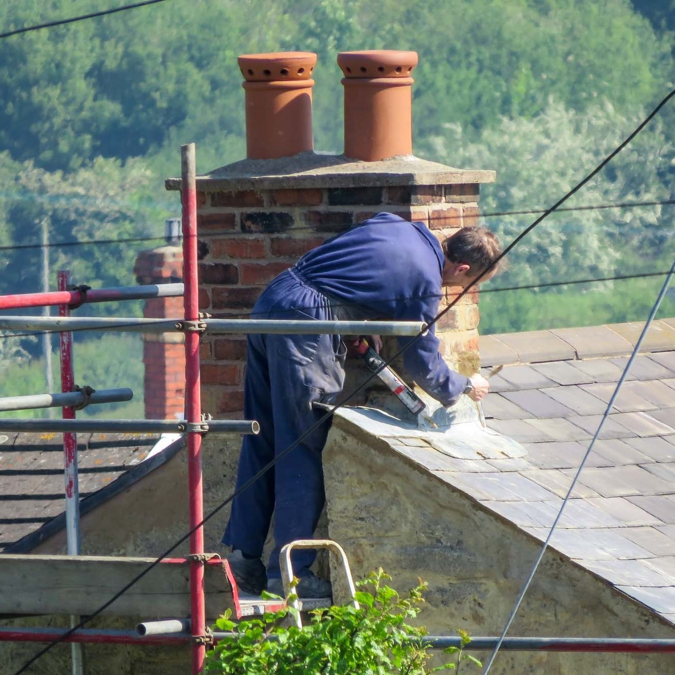 Roofer repairing flashing on chimney stack on a slate roof of a domestic house. Standing on scaffolding. Close up, high angle point of view. Countryside background.